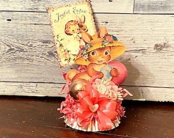 May include: A vintage Easter decoration featuring a bunny wearing a hat and holding an egg. The decoration includes a card that reads "Joyful Easter", pink ribbons, and a decorative base.