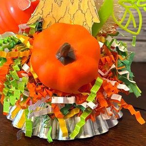 May include: A small, orange pumpkin sits on top of a silver cupcake liner filled with colorful shredded paper. The pumpkin has a brown stem and is surrounded by green, orange, and white paper shreds.