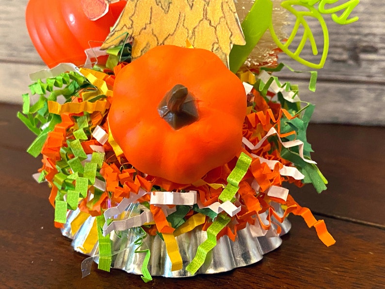 May include: A small, orange pumpkin sits on top of a silver cupcake liner filled with colorful shredded paper. The pumpkin has a brown stem and is surrounded by green, orange, and white paper shreds.