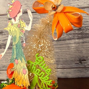 May include: A Thanksgiving-themed tabletop decoration featuring a paper doll wearing a green dress with orange accents, a small artificial tree with a white bristle brush, and a green sign that reads "Give Thanks".