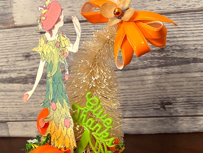 May include: A Thanksgiving-themed tabletop decoration featuring a paper doll wearing a green dress with orange accents, a small artificial tree with a white bristle brush, and a green sign that reads "Give Thanks".