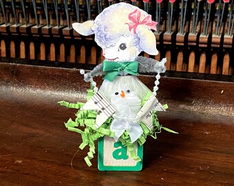 May include: A white lamb figurine with a green bow and a pink bow on its head, holding a string of white beads. The lamb is sitting on a green block with the letter 'a' on it. The block is surrounded by green and white shredded paper and a white bird figurine with a red beak.