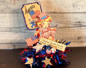 May include: A patriotic centerpiece featuring a vintage illustration of a girl in a red and white striped dress and hat, holding an American flag. The base is decorated with red, white, and blue tinsel, stars, and small flags. Text reads "Yankee Doodle Dandy" and "America."