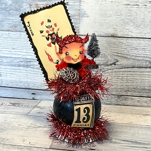 May include: A black and red Halloween decoration featuring a devil character with red horns and a red dress, holding a playing card with the word "Joker" on it. The decoration is surrounded by red tinsel and a black orb with a calendar that reads "Today is 13 Friday".