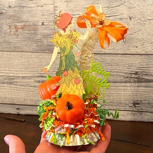 May include: A Thanksgiving-themed centerpiece featuring a paper doll dressed in a green and yellow dress with a red hat, surrounded by orange pumpkins, green and orange shredded paper, and a gold and orange ribbon. The centerpiece is on a silver cupcake liner and says "Give Thanks".
