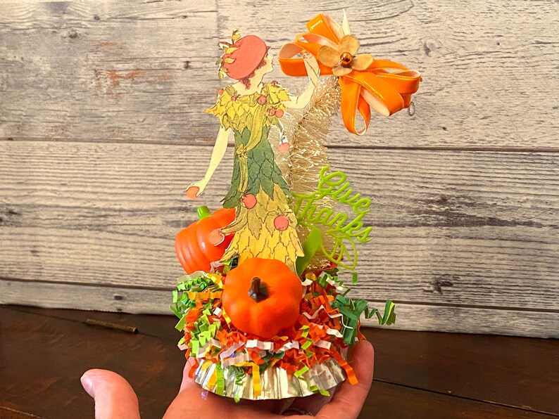May include: A Thanksgiving-themed centerpiece featuring a paper doll dressed in a green and yellow dress with a red hat, surrounded by orange pumpkins, green and orange shredded paper, and a gold and orange ribbon. The centerpiece is on a silver cupcake liner and says "Give Thanks".