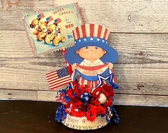 May include: A patriotic centerpiece featuring a child holding a banner that says "America" and wearing a red, white, and blue hat. The centerpiece is decorated with red, white, and blue streamers, stars, and a small American flag.