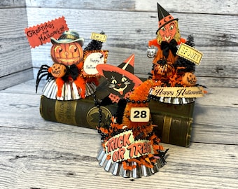 May include: Three Halloween-themed decorations made from vintage tin pie plates. The decorations feature a jack-o'-lantern, a black cat, and a witch. Each decoration is adorned with orange and black paper, ribbon, and other embellishments. The decorations are sitting on top of an old book.