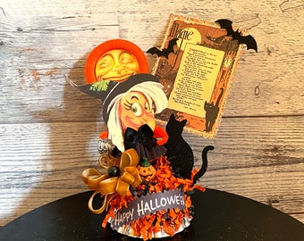 May include: A Halloween themed centerpiece featuring a witch figurine, a black cat, a pumpkin, and a paper sign with a poem about magic. The centerpiece is decorated with orange and black tissue paper and a gold bow.