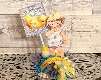 May include: A vintage Easter centerpiece featuring a blue and white paper doll holding a finger to her lips, a yellow chick, and a blue and white paper sign that reads "New South Wales". The centerpiece is decorated with blue and yellow flowers and a yellow ribbon.