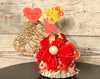 May include: A Valentine's Day themed craft decoration featuring a red heart with the text "A Valentine for YOU!" and a cutout wooden heart with the word "LOVE" in a decorative font. The decoration also includes a red bow, a white pearl, and a red and white paper cupcake liner.