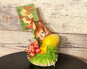 May include: A vintage Easter centerpiece featuring a brown bunny figurine holding a yellow egg, a pink bow, and a green card with the text "Happy Hoppy Easter".