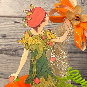 May include: A paper doll wearing a green dress with a red hat and holding a pumpkin. The doll is standing in front of a white and gold tinsel garland and an orange ribbon bow with a brown flower.