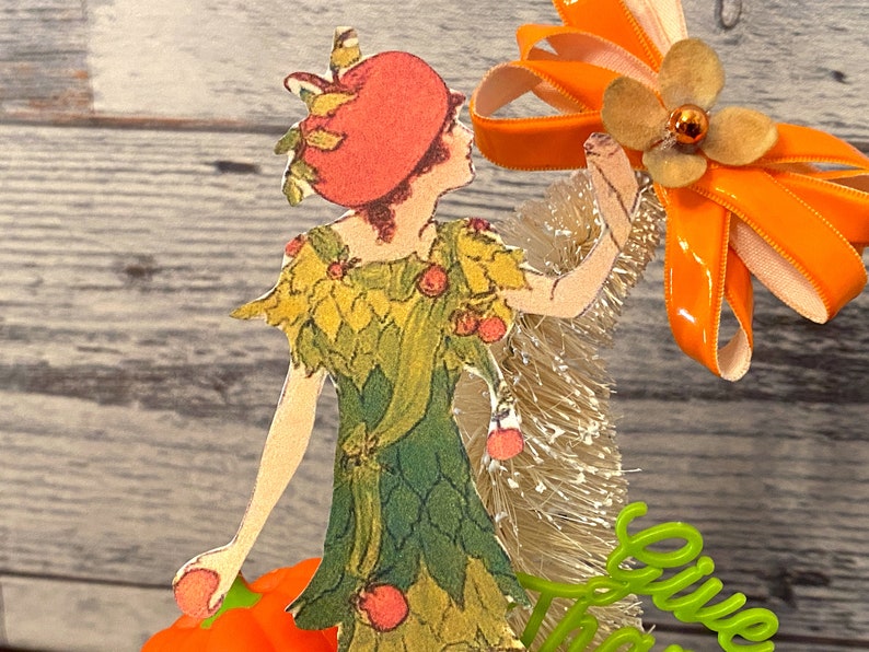 May include: A paper doll wearing a green dress with a red hat and holding a pumpkin. The doll is standing in front of a white and gold tinsel garland and an orange ribbon bow with a brown flower.