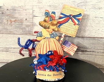 May include: A patriotic figurine featuring a vintage illustration of a girl in a red, white, and blue dress. The base is decorated with stars, confetti, and the text "America the Beautiful". Includes an American flag and a sign.