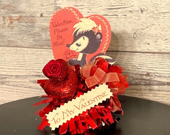 May include: A red glitter heart with a red rose on top, surrounded by red tinsel and a red ribbon. A vintage Valentine's Day card with a cartoon skunk holding a rose and the text "Valentine, Please Be Mine!" is attached to the top of the heart. A small tag with the text "To My Valentine" is attached to the bottom of the heart.