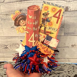 May include: A patriotic Fourth of July decoration featuring a red, white, and blue color scheme. The centerpiece includes a decorative pillar with the text "Will Go off on the 4th". Additional elements include a vintage illustration, a miniature American flag, and star-shaped embellishments.