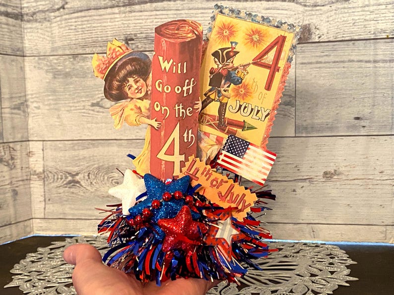 May include: A patriotic Fourth of July decoration featuring a red, white, and blue color scheme. The centerpiece includes a decorative pillar with the text "Will Go off on the 4th". Additional elements include a vintage illustration, a miniature American flag, and star-shaped embellishments.