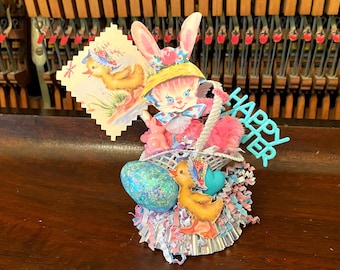 May include: An Easter-themed decorative arrangement featuring a pink bunny with a basket, holding a card with a duck illustration. Includes a glittery blue egg, a yellow duck, and the words "HAPPY EASTER".
