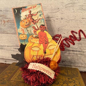 May include: A decorative Halloween centerpiece featuring a vintage-style card with illustrations of devils and jack-o'-lanterns, a die-cut orange pumpkin with a seated red devil figure, red metallic tinsel, a small plastic fork, and a sign reading Halloween.