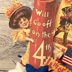 May include: Vintage patriotic Fourth of July decoration featuring a smiling girl, a soldier, and an American flag. The red pillar reads "Will Go off on the 4th". Includes a blue bow and star.