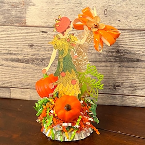 May include: A vintage-style paper doll figurine dressed in a green and yellow dress with a red hat, holding a small orange pumpkin. The doll is surrounded by orange and green shredded paper and a large orange bow. The text "Give Thanks" is written in green letters on the side of the doll.