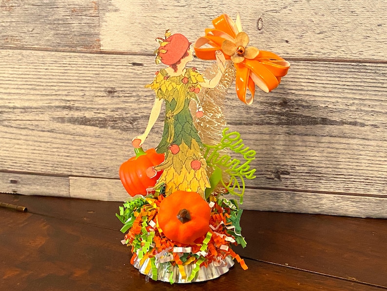 May include: A vintage-style paper doll figurine dressed in a green and yellow dress with a red hat, holding a small orange pumpkin. The doll is surrounded by orange and green shredded paper and a large orange bow. The text "Give Thanks" is written in green letters on the side of the doll.