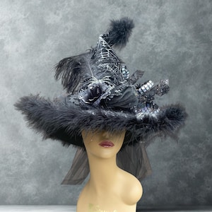 May include: A black witch hat featuring a wide brim trimmed with black feathers, a pointed crown with a silver spiderweb pattern, a black rose accent, dark tulle fabric, and a black pom-pom at the top, displayed on a beige mannequin head.