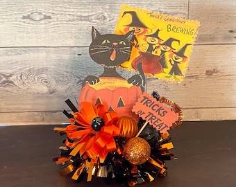 May include: A Halloween-themed decoration featuring a black cat emerging from a carved pumpkin. A sign reads "Tricks or Treat" and a card with witches and the text "There's Something Brewing" is in the background. The decoration includes orange and black accents.