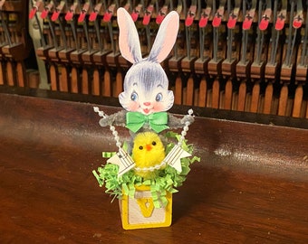 May include: A yellow chick with a green bow sits in a yellow block with the letter V on it. A gray bunny with a green bow is perched on top of the chick. The bunny and chick are surrounded by green and white paper shreds.