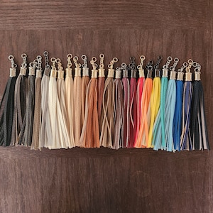 May include: A collection of 20 colourful leather tassels with gold metal rings. The tassels are arranged in a row and are all different colours, including red, blue, green, yellow, and brown.