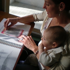May include: A woman is reading a book to a young child. The book is open to a page with a pink and white patterned design. The woman is wearing a white sweater and the child is wearing a white shirt.