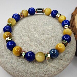 May include: A beaded bracelet featuring alternating blue and yellow spherical beads. The bracelet is accented with small, metallic spacers. The beads are arranged in a circular pattern, resting on a gray stone surface.