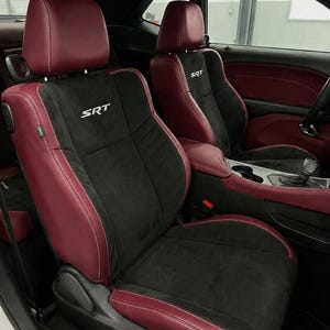 May include: Two automotive bucket seats featuring a combination of deep red leather and black suede-like material. The backrests display the white embroidered text SRT. The seats include adjustable headrests and visible white contrast stitching.
