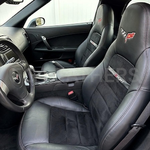 May include: Black leather car seats with suede inserts. The seats have red stitching and the Corvette logo. The headrests feature the Grand Sport logo and a checkered flag emblem. The interior includes a steering wheel and dashboard.