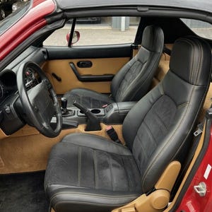 May include: The interior of a red convertible car featuring black leather bucket seats with tan stitching, a black steering wheel, a manual gear shifter, and a tan carpeted floor. A black floor mat with the white text MX-5 is visible on the driver side.