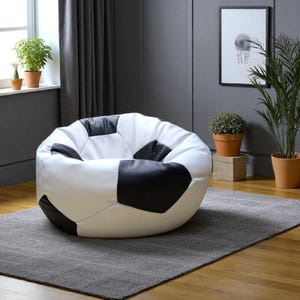 Puede incluir: Un gran puf con forma de balón de fútbol. La silla es blanca con paneles negros en forma de pentágono. Se encuentra sobre una alfombra gris en una habitación con ventana, plantas y un cuadro enmarcado.