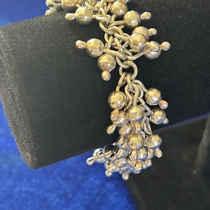 Könnte beinhalten: Ein silberfarbenes Bettelarmband mit einer dichten Ansammlung kleiner, runder Metallperlen, die an einer Gliederkette befestigt sind. Die Perlen haben eine glatte, reflektierende Oberfläche und sind in mehreren Schichten am Armband angeordnet.
