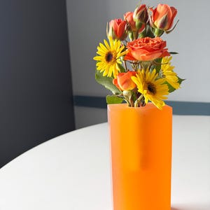 Könnte beinhalten: Ein Strauß orangefarbener und gelber Blumen in einer leuchtend orangenen Vase. Die Vase ist aus Glas und hat eine quadratische Form.
