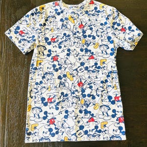 May include: White t-shirt featuring a repeating pattern of Mickey Mouse in various poses. The cartoon character is depicted in blue, red, yellow, and black on a white background. The short-sleeved shirt has a crew neck.