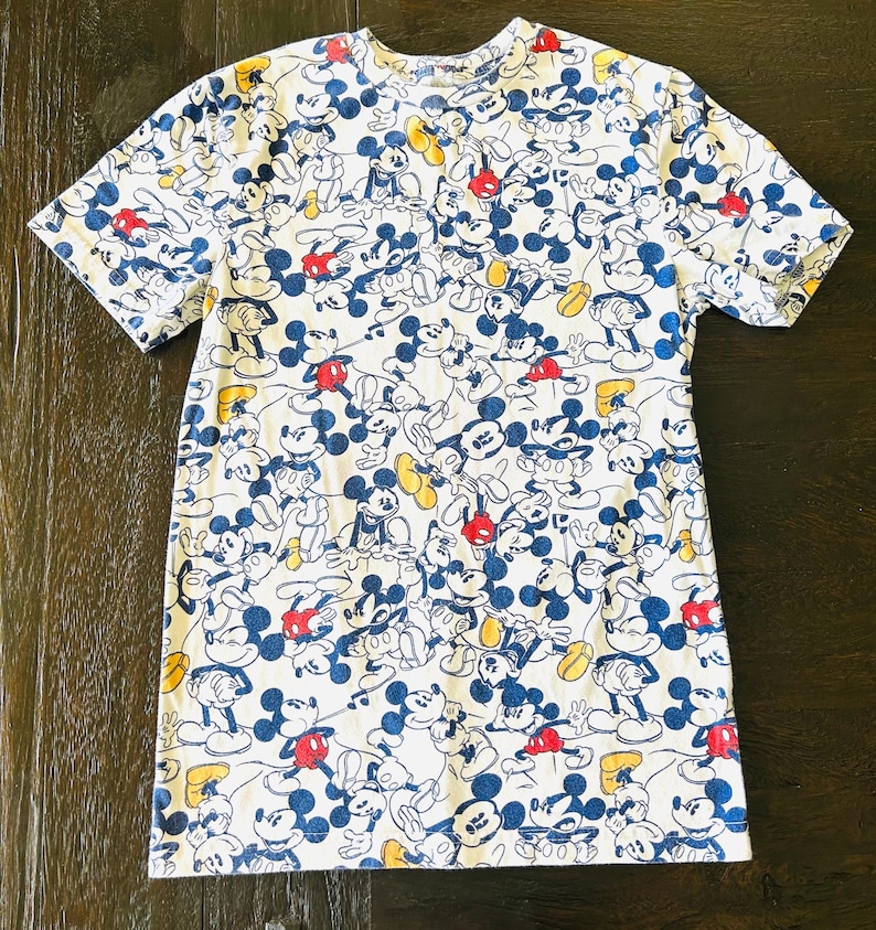 May include: White t-shirt featuring a repeating pattern of Mickey Mouse in various poses. The cartoon character is depicted in blue, red, yellow, and black on a white background. The short-sleeved shirt has a crew neck.
