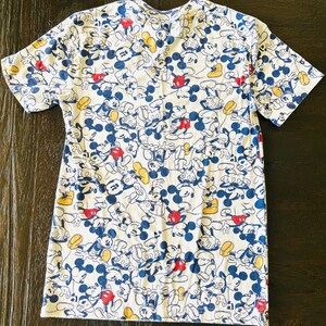 May include: White t-shirt featuring a repeating pattern of Mickey Mouse in various poses. The design includes blue, red, and yellow accents on a white background. The shirt has short sleeves and a crew neck.