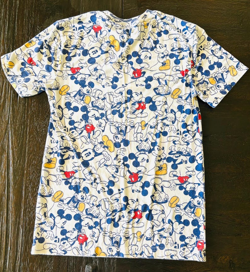 May include: White t-shirt featuring a repeating pattern of Mickey Mouse in various poses. The design includes blue, red, and yellow accents on a white background. The shirt has short sleeves and a crew neck.