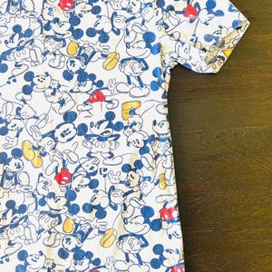 May include: White t-shirt featuring a repeating pattern of Mickey Mouse in various poses. The cartoon character is depicted with blue, red, and yellow accents. The shirt has short sleeves and a crew neck.