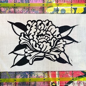 Puede incluir: Un trozo rectangular de tela color hueso con un estampado en bloque de tinta negra de una flor estilizada con pétalos superpuestos y hojas puntiagudas. Una regla de plástico amarilla es visible a lo largo de los bordes superior e inferior.