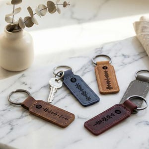 May include: A collection of keychains in various colors, including brown, blue, tan, gray, and burgundy. Each keychain features a metal ring and a rectangular leather tag with a unique audio waveform design. A key is attached to one keychain.