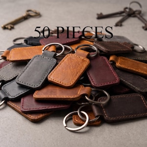 May include: A collection of 50 rectangular leather keychains in various shades of brown, black, and burgundy, each featuring a metal split ring. The text 50 PIECES is displayed above the pile of keychains, which are scattered alongside several antique metal keys.