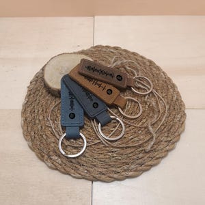 May include: Four leather keychains with a unique design featuring a sound wave pattern and a small circle. The keychains are brown and blue and are displayed on a woven straw mat.