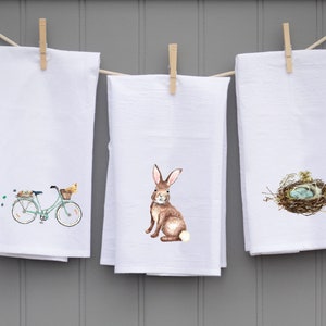 May include: Three white kitchen towels with different Easter-themed designs. The first towel features a green bicycle with a basket of flowers and a chick. The second towel features a brown rabbit. The third towel features a bird's nest with blue eggs.