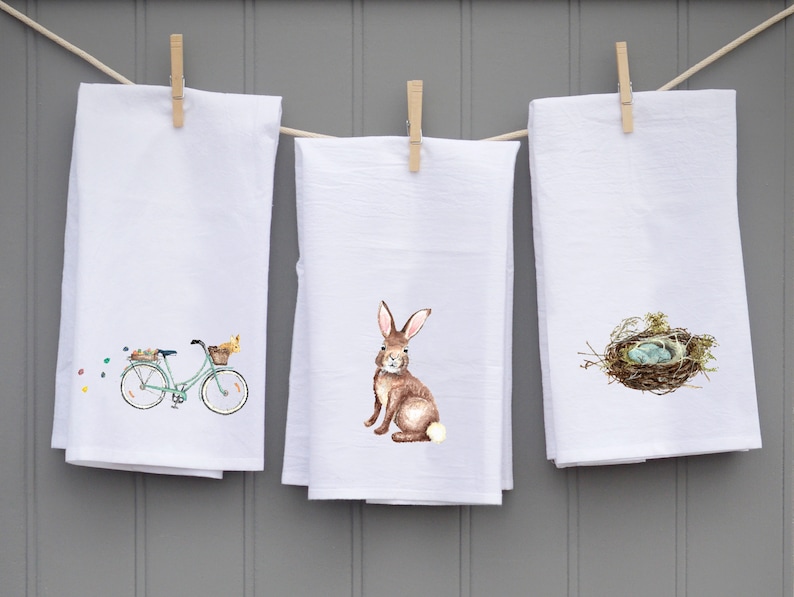 May include: Three white kitchen towels with different Easter-themed designs. The first towel features a green bicycle with a basket of flowers and a chick. The second towel features a brown rabbit. The third towel features a bird's nest with blue eggs.